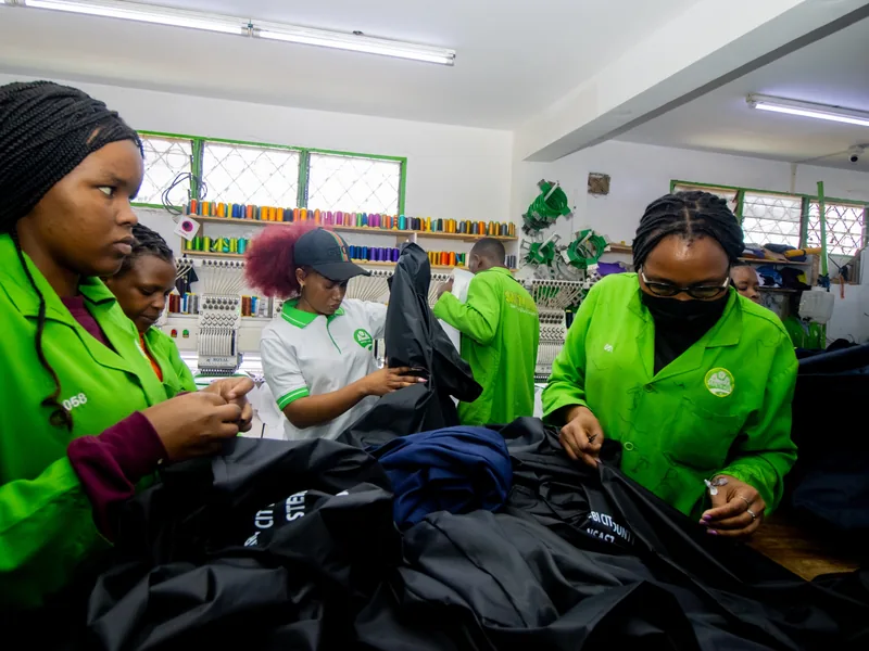 Safety Hub production team in green coats sewing dark workwear in front of multi-head embroidery machines in the Nairobi workshop
